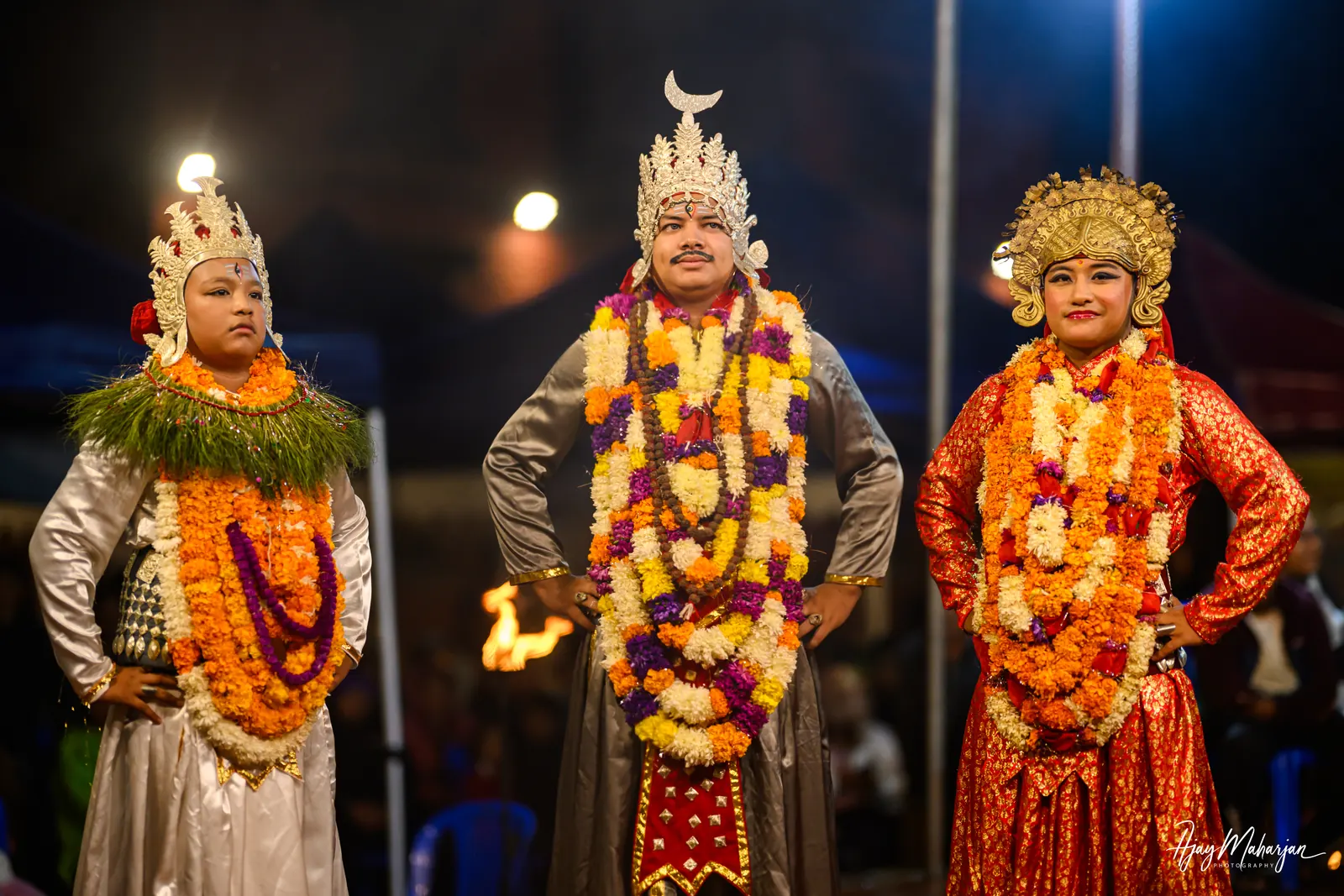 Three crowned performers in garlands stand together at night