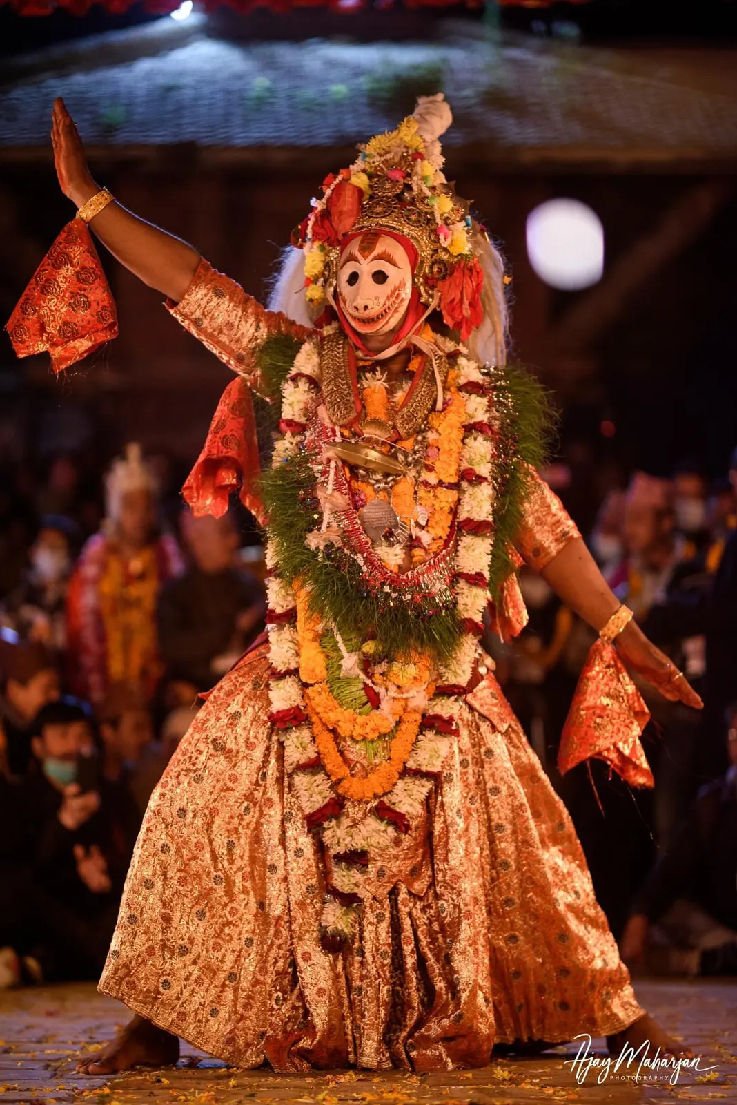 A dancer in a white monkey mask, flower crown and gold robes strikes a wide pose