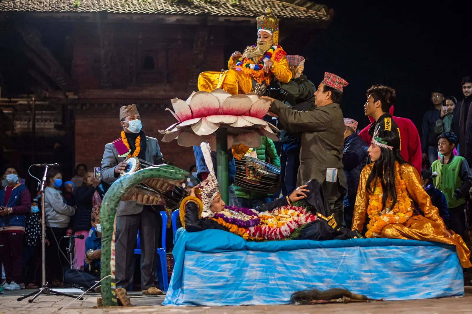 Actors perform a scene on a raised stage with a large painted serpent, in front of the crowd