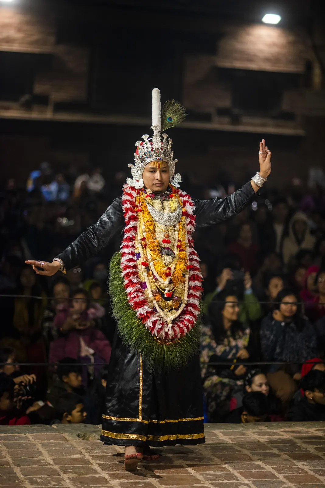 A dancer in a tall silver crown and garland raises one arm